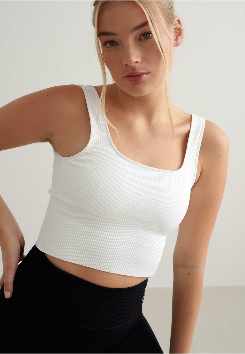 Bílý ribovaný crop top se čtvercovým výstřihem a silnými ramínky, spárovaný s černými legínami s vysokým pasem, předvádějící kontrastní barvy a textury.