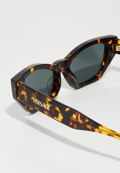 Schildkrötensonnenbrille mit dunklen Gläsern; dicke, eckige Rahmen mit goldfarbenem Logo-Detail an den Schläfen. Die strukturierte Oberfläche verstärkt das Design.