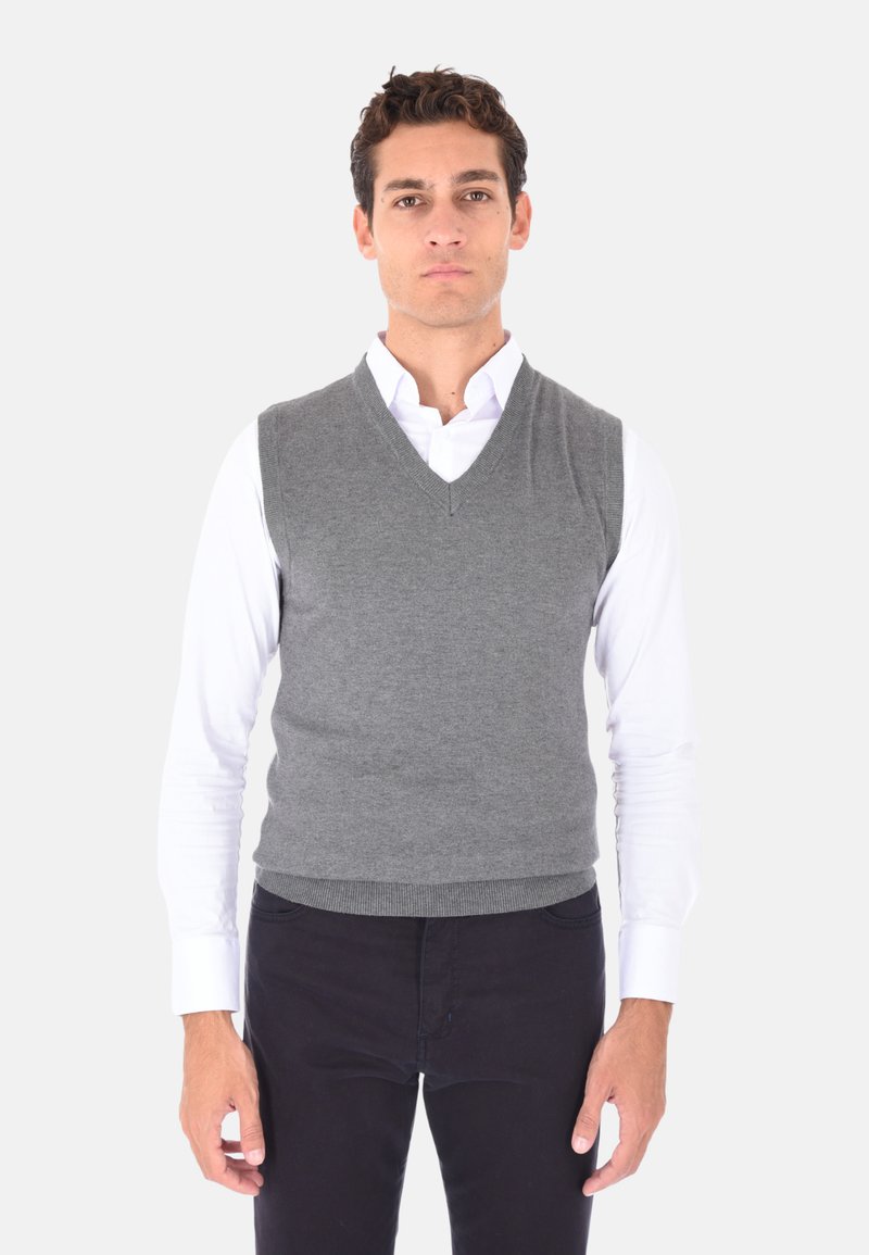 Maglione grigio senza maniche con scollo a V. Realizzato in morbido tessuto a maglia, presenta una silhouette aderente e un orlo a costine. Indossato sopra una camicia bianca.