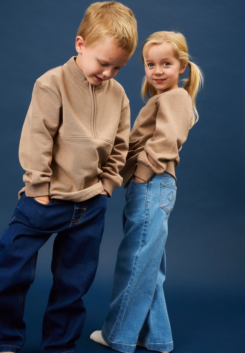 Deux jeunes enfants debout devant un fond bleu, portant des sweat-shirts beiges et des jeans bleus, les mains dans les poches.