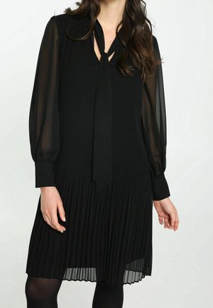 Frau trägt ein schwarzes Kleid mit transparenten langen Ärmeln, einer Halsbinde und einem plissierten Rock, steht vor einem einfarbigen Hintergrund.