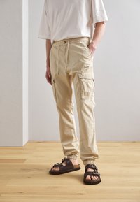 Pantaloni cargo beige con polsini elastici, vita con cordoncino e tasche laterali. Indossati con sandali marroni con dettagli a fibbia.