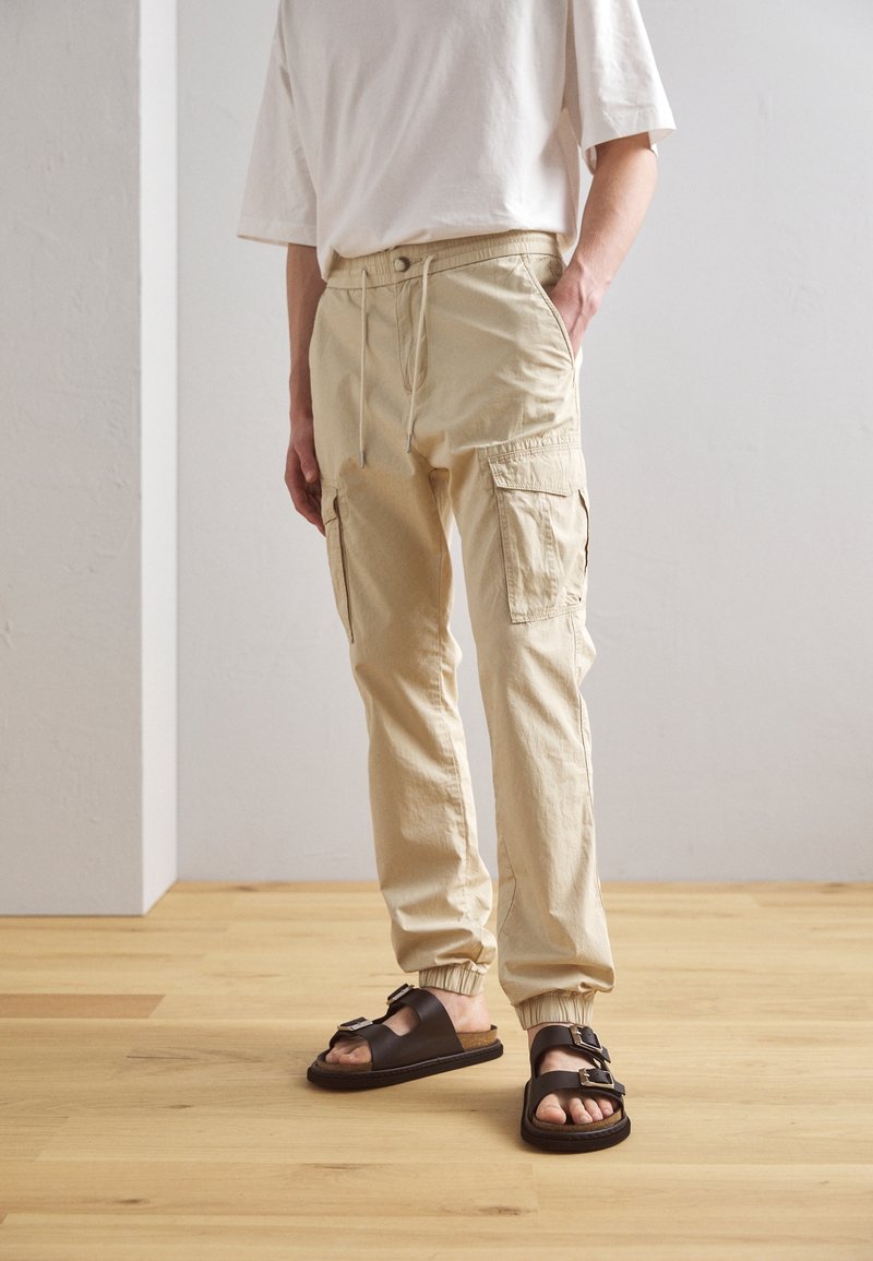 Pantaloni cargo beige con polsini elastici, vita con cordoncino e tasche laterali. Indossati con sandali marroni con dettagli a fibbia.