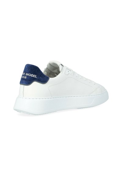 Witte low-top sneakers met een blauwe suède hieltab, voorzien van witte geborduurde tekst, vetersluiting aan de voorkant en een dikke witte rubberen zool.