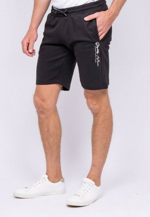 Homme portant un short noir décontracté avec un texte blanc sur la jambe gauche et des baskets blanches, debout, la main droite dans la poche.