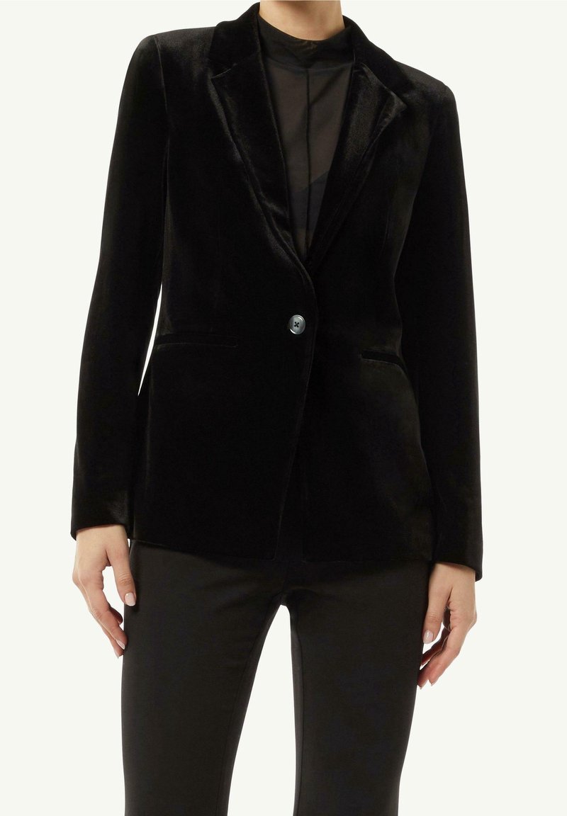 Blazer en velours noir à un bouton porté sur un top noir transparent et un pantalon noir adapté, modèle montré du cou aux hanches.