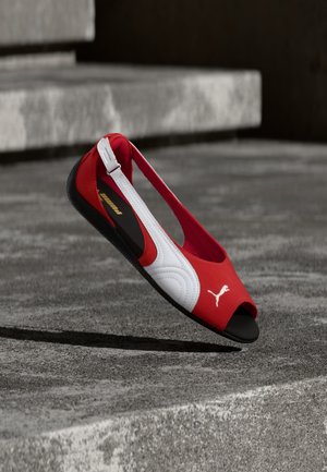 Zapato plano abierto en la puntera con tiras traseras de Puma en rojo y blanco flotando sobre escalones de hormigón con suela negra y logo de Puma en la parte delantera.