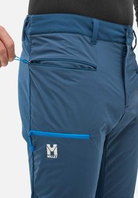 Main masculine tirant sur la fermeture éclair d'une poche de pantalon Millet bleu pour l'extérieur, montrant la texture du tissu et le logo sur la poche de cuisse.