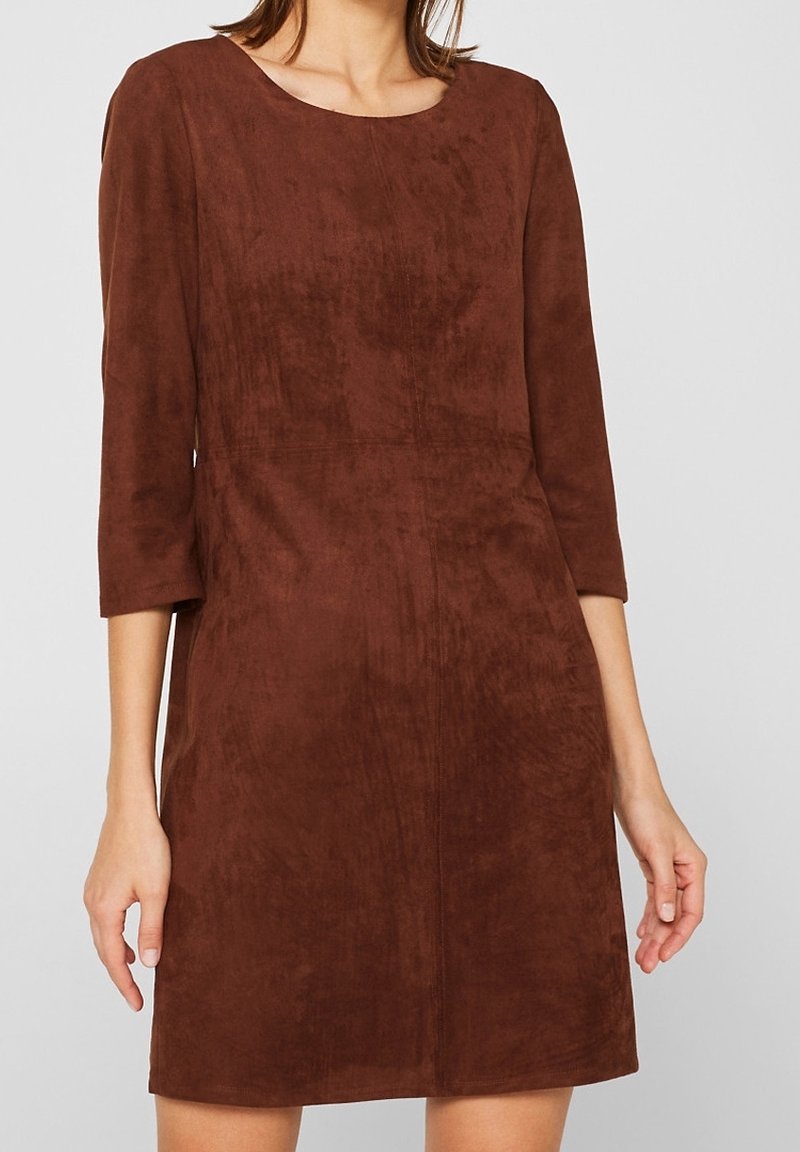 Femme portant une robe en daim marron avec des manches trois-quarts et un col rond, debout devant un fond clair uni.