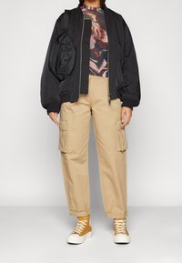 Czarna kurtka typu bomber, kolorowy top w wzory, beżowe spodnie cargo z kieszeniami bocznymi i żółte sneakersy za kostkę. Prosta czarna torba na ramię.