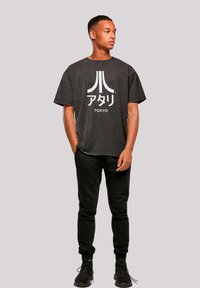 Schwarzes oversized T-Shirt aus Baumwolle mit einem retro Atari-Logo in Weiß, kombiniert mit schwarzen Jogginghosen und schwarzen Sportschuhen.