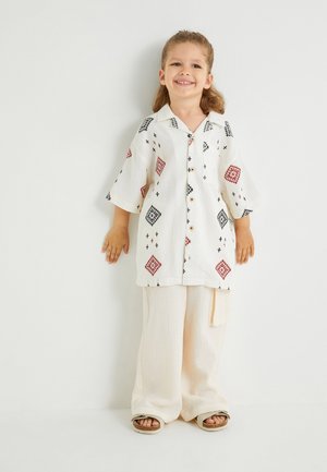 Camisa blanca de manga corta con patrones de diamantes rojos y negros, con botones en el frente. Pantalones anchos marfil. Niño de pie contra un fondo neutro.
