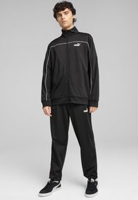 Chándal deportivo negro con piping blanco, chaqueta con cremallera y pantalones con cintura elástica. Presenta el logo de Puma en la chaqueta y los pantalones. Tela texturizada.