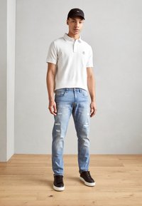 Hvit polo skjorte med korte ermer og krage, kombinert med lys blå slitte jeans og svarte sneakers, mot en lys bakgrunn.