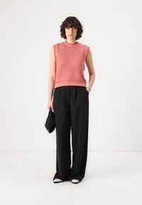 Rotes ärmelloses Stricktop mit strukturiertem Muster und gerippten Kanten, kombiniert mit hoch taillierten schwarzen weit geschnittenen Hosen und einer schwarzen Handtasche.