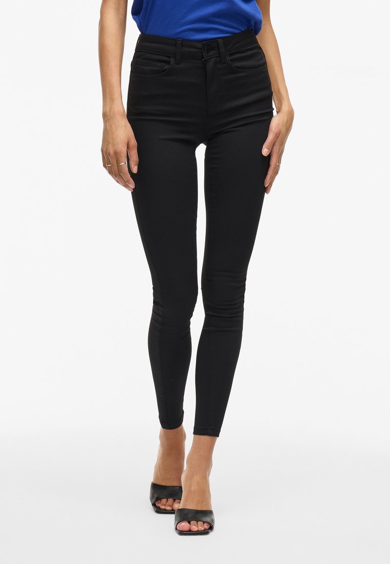 Femme portant un jean skinny taille haute noir, un haut bleu et des sandales à talons noires à bout ouvert, debout sur fond blanc.