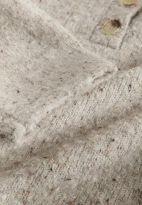 Béžová vlnená tkanina s textúrovaným povrchom, obsahujúca malé fliačiky a dva veľké okrúhle gombíky pozdĺž okraja.