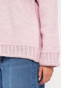 Różowy sweter dzianinowy z teksturowanym, prążkowanym dołem i mankietami. Luźny krój z miękkiego, lekkiego materiału, noszony na niebieskie dżinsy.
