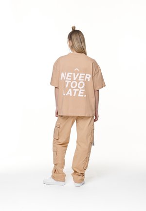 T-shirt oversize beige chiaro con testo bianco che recita "NEVER TOO LATE." abbinato a pantaloni cargo beige larghi e sneakers bianche.