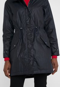 Longue veste noire avec cordons à la taille et poches à rabat, portée sur un haut rouge à manches longues, montrée sur une personne des épaules aux cuisses.