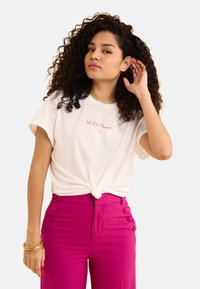 Camiseta de algodón blanca con el texto "Corazón Salvaje" en rosa, anudada en la cintura. Combinada con pantalones fucsias de talle alto con detalles de botones.