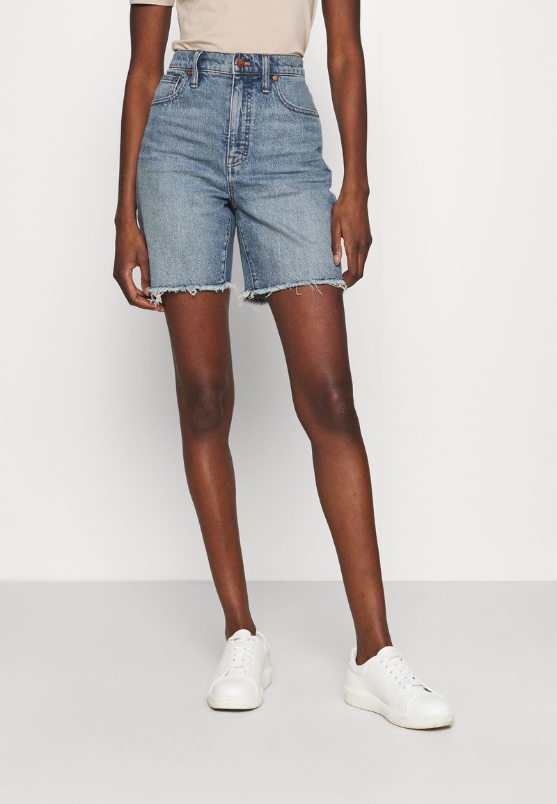 Blå denimshorts med hög midja och fransad fåll, i kombination med vita sneakers. Slät textur och klassisk femfickorsdesign.