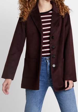 Mujer vestida con un abrigo marrón oscuro abotonado sobre una camisa de rayas rojas, blancas y negras, con jeans azules, de pie contra un fondo liso.