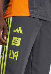 Grå sportbukser med limegrønne accenter og logoer, der har en glat tekstur og sidelstænger. Materialet virker let og åndbart.