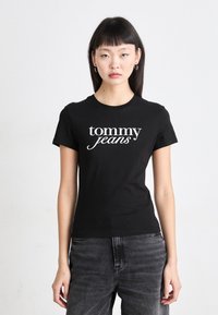 Svart bomulls T-skjorte med hvit "tommy jeans" logo, kort ermer og rund hals, kombinert med mørk grå bukser.