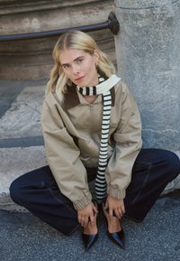 Veste bomber beige avec un col en velours côtelé, associée à une écharpe rayée noir et blanc. Jean foncé et talons noirs pointus. Fond texturé.