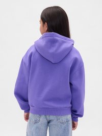 Bambino con lunghi capelli scuri che indossa un ampio hoodie viola e jeans azzurri chiari, di spalle contro uno sfondo chiaro e semplice.