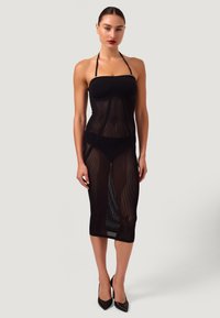 Donna con capelli raccolti che indossa un vestito in rete nero con collo ad anello sopra biancheria intima nera e tacchi neri, in piedi davanti a uno sfondo neutro.