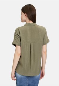 Olijfgroene kortekraag shirt met korte mouwen, voorzien van een plooi-detail aan de achterkant en een afgeronde zoom. De stof lijkt lichtgewicht en gestructureerd.