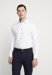 Camicia bianca a maniche lunghe con bottoni blu, colletto strutturato e design aderente, abbinata a pantaloni scuri e una cintura nera.