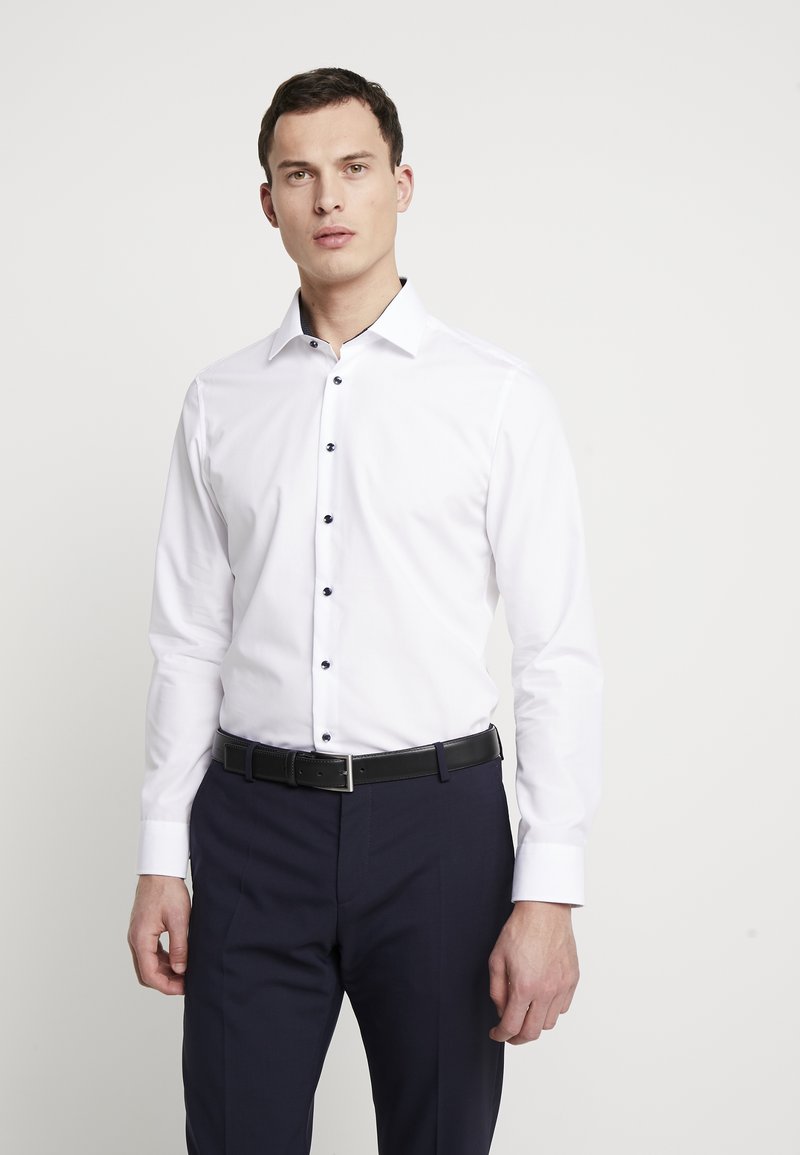 Camicia bianca a maniche lunghe con bottoni blu, colletto strutturato e design aderente, abbinata a pantaloni scuri e una cintura nera.