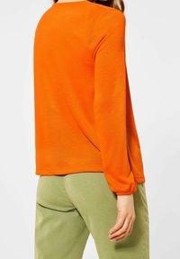 Dlouhý oranžový svetr s kulatým výstřihem, měkkou texturou a elastickými manžetami, kombinovaný se světle zelenými kalhotami.