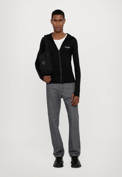 Svart hettejakke med "SILENCE" tekst, lange ermer og tilpasset design, sammen med grå rette jeans og svarte sko.