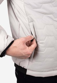 Chaqueta acolchada gris claro con patrón acolchado hexagonal, bolsillo lateral con cremallera y cintura elástica. La tela es lisa y ligeramente brillante.