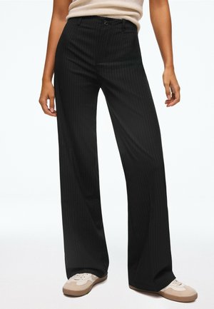 Une femme porte un pull à col roulé crème à manches courtes, un pantalon noir à jambes larges et des baskets beiges sur un fond blanc uni.