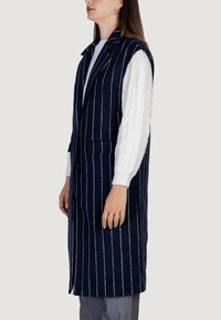 Námořnická modrá bezrukávová vesta s bílými vertikálními pruhy, vyrobená z texturované tkaniny. Obsahuje klopy a dvě přední kapsy.