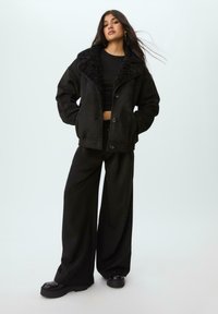 Crna jakna od lažne suede tkanine s teksturiranim crnim krznom oko vrata, oversized kroj, te prednjim dugmadima; uparena s širokim crnim hlačama i platformama.