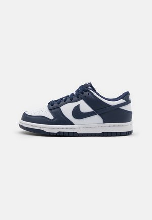 Low-top Nike Dunk sneaker i hvid og navyblåt læder med navyblå snørebånd og hvid mellemsål på en ensfarvet baggrund.