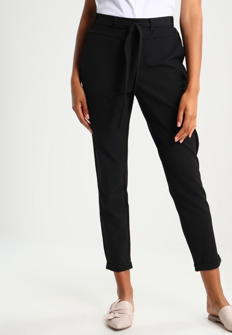 Pantalons noir sur mesure avec une ceinture à nouer, coupe slim, tissu lisse et poignets retroussés. Portés avec des chaussures décontractées de couleur claire.