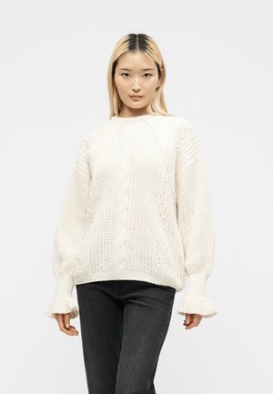 Femme aux longs cheveux blonds portant un pull blanc à torsades avec des poignets évasés et un pantalon noir, debout devant un fond clair uni.