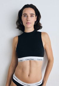 Crop top sans manches noir avec un col montant et une bande élastique blanche portant le logo "Calvin Klein" en bas. Texture lisse.