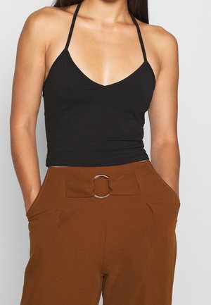 Vrouw draagt een zwart halter crop top en een hooggetailleerde broek in bruin met een stoffen riem en een metalen ring, handen in de zakken, tegen een effen achtergrond.