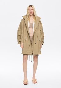 Beżowa, oversize parka z regulowanym w pasie sznurkiem, przednim zamkiem błyskawicznym i dwiema dużymi kieszeniami. Nosi się ją na beżowy top crop i spódnicę.
