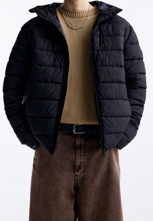 Persona que lleva una chaqueta acolchada negra sobre un suéter de punto beige, pantalones marrones y un collar de cadena plateada, de pie frente a un fondo blanco.