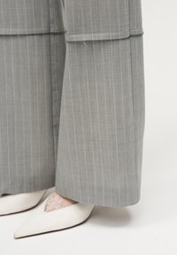 Pantalon large gris à fines rayures associé à des talons blancs à bouts pointus partiellement recouverts de chaussettes en dentelle, visible de la partie inférieure du mollet.