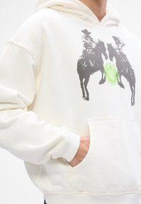 Hooded džemperis gaiši krēmkrāsā, ar grafiku, kurā attēloti divi jātnieki uz zirgiem, zaļš aplis logotips un priekšējā kabata. Tekstūrēta auduma.